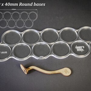 May include: Clear acrylic base holder for 10 x 40mm round bases, with a wooden handle. The holder has a wavy design with ten circular cutouts. The text "For 10 x 40mm Round bases" is at the top. The holder is 24cm long.
