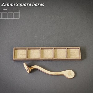 Puede incluir: Una base de madera para 5 bases cuadradas, cada una de 25 mm. La base es rectangular con cinco recortes cuadrados. Tambi&eacute;n se incluye un asa de madera curvada. El texto "For 5 - 25mm Square bases" est&aacute; en la parte superior.