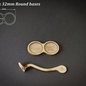 May include: Two wooden round bases, each 32mm in diameter, with a connecting handle. The bases are designed to hold miniatures. The text "For 2 x 32mm Round bases" is at the top. The text "Jimmy's Minis" is visible on one base.