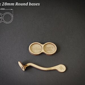 May include: Two wooden round bases for miniatures, each with a 28mm diameter, and a curved wooden handle. The text "For 2 x 28mm Round bases" is at the top. The handle has a small circular base.