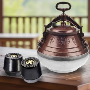 以下が含まれることがあります： ハンドルと2つの黒い円錐形のアクセサリーが付いた、銅色の圧力鍋。鍋は銀色のベースと装飾的な花の模様があります。アクセサリーには「Rashko Baba Ltd」の文字が入っています。木製のテーブルの上に置かれたアイテムです。