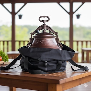 Puede incluir: Una olla a presión de color cobre con una bolsa de transporte negra está sobre una mesa de madera. La olla a presión tiene un asa y patrones florales decorativos. La bolsa tiene correas ajustables y una hebilla.