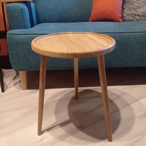 Puede incluir: Pequeña mesa auxiliar redonda de madera clara con borde elevado. Tiene tres patas anguladas y un acabado de madera natural. Mide unos 50 cm de diámetro y 46 cm de altura.