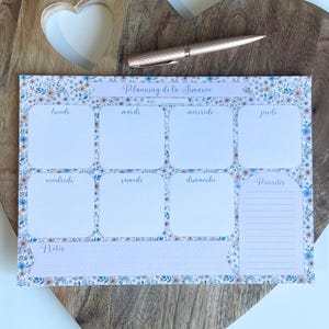 Peut inclure: Planificateur hebdomadaire avec un motif floral bleu, blanc et marron. Le planificateur comprend des sections pour chaque jour de la semaine, une section notes et une section priorités. Un stylo or rose repose sur une surface en bois.