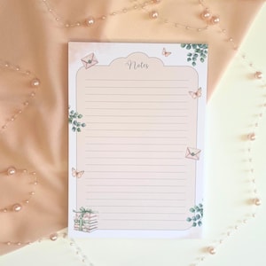 Peut inclure: Un bloc-notes rose pâle avec des pages lignées et des éléments décoratifs. Le bloc-notes porte le mot "Notes" en haut, avec des illustrations de papillons, d'enveloppes et de verdure. Une pile de cadeaux est en bas.