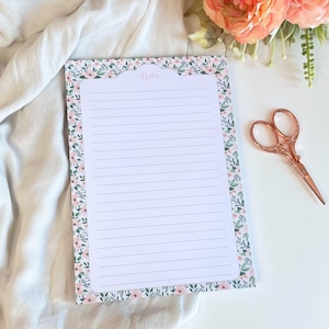 Peut inclure: Bloc-notes à motifs floraux avec le mot "Notes" en haut. Le bloc-notes a une bordure de fleurs roses et de feuilles vertes. Des ciseaux en or rose sont posés à droite.