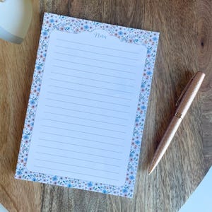 Può includere: Un blocco note a fantasia floreale con pagine a righe e la parola "Notes" in alto. Una penna color oro rosa è appoggiata accanto al blocco note su una superficie di legno. Il bordo del blocco note presenta piccoli fiori blu e viola.