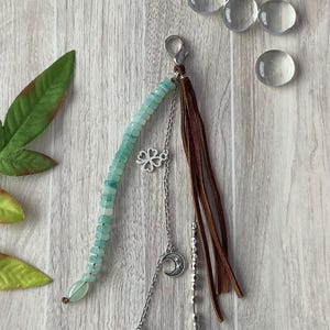 May include: A decorative charm featuring a strand of light green beads, a silver chain with a four-leaf clover and crescent moon charm, and a cluster of brown suede leather strips. The accessory is attached to a silver lobster claw clasp.