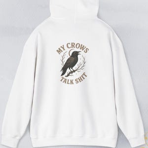 Könnte beinhalten: Weißer Kapuzenpullover mit dem Text "MY CROWS TALK SHIT" in einem kreisförmigen Design. Das Design zeigt eine schwarze Krähe, die auf einem Ast sitzt. Der Kapuzenpullover hat eine Kapuze mit Kordelzug und eine Kängurutasche.