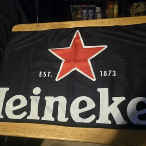 May include: A black rectangular fabric banner featuring a large red five-pointed star centred above the white text Heineken. The text EST. 1873 is printed in white on either side of the star. A registered trademark symbol appears at the end of the word.