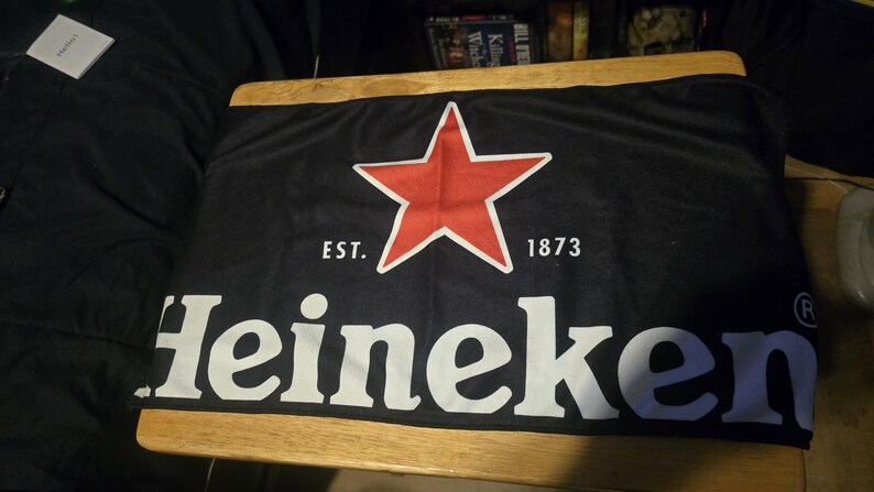 May include: A black rectangular fabric banner featuring a large red five-pointed star centred above the white text Heineken. The text EST. 1873 is printed in white on either side of the star. A registered trademark symbol appears at the end of the word.