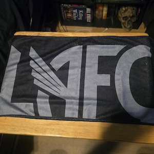 May include: A rectangular black sports scarf featuring the letters LAFC in large, bold, grey block lettering. The letter A is stylised with a wing design integrated into its structure. The fabric has a smooth, uniform texture throughout the surface.