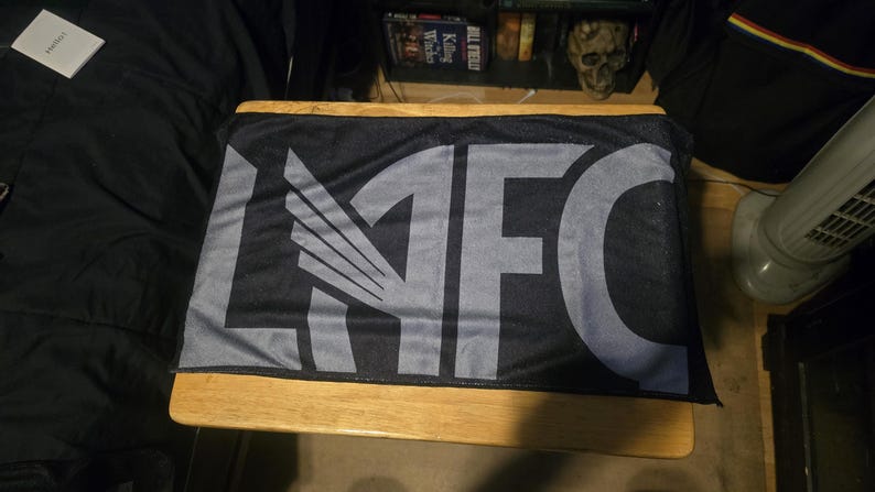 May include: A rectangular black sports scarf featuring the letters LAFC in large, bold, grey block lettering. The letter A is stylised with a wing design integrated into its structure. The fabric has a smooth, uniform texture throughout the surface.