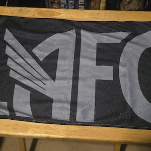 May include: A rectangular black sports scarf featuring the letters LAFC printed in a large, bold, grey font across the centre. The letter A is stylised with a wing design integrated into its structure, creating a distinct graphic appearance.