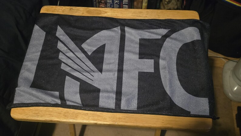 May include: A rectangular black sports scarf featuring the letters LAFC printed in a large, bold, grey font across the centre. The letter A is stylised with a wing design integrated into its structure, creating a distinct graphic appearance.