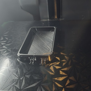 May include: A rectangular black 3D printed object with a hollow center and visible layer lines, resting on a textured build plate. The object features a thin frame with small protruding supports along one side, showing the printing process in progress.
