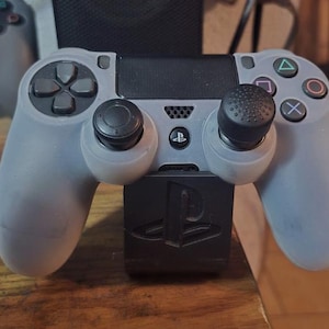 May include: A grey PlayStation 4 controller with black joysticks and buttons, on a black charging dock. The controller has the standard button layout with a directional pad, action buttons, and a touchpad. The charging dock features the PlayStation logo.