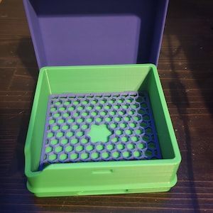May include: A 3D-printed storage box with a removable tray. The box is composed of a green base and a purple lid. The tray is green with a honeycomb pattern and a purple border. The box is open, revealing the interior.