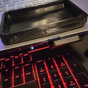 May include: A black, rectangular, 3D-printed phone case with a hollow center and side cutouts for buttons. The case is resting on a laptop keyboard that features black keys with red backlighting and a screen displaying the text the building's.