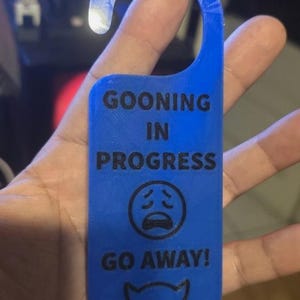 Könnte beinhalten: Ein blauer Türhänger mit schwarzem Text "GOONING IN PROGRESS" und "GO AWAY!". Er zeigt ein trauriges Gesicht und ein Teufelsgesicht. Die Oberseite hat einen gebogenen Griff zum Aufhängen.