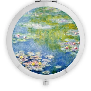 May include: A round, silver compact mirror featuring a vibrant impressionist painting of water lilies in a pond. The artwork showcases a palette of blues, greens, yellows, and pinks, with the lilies and their reflections creating a serene scene.