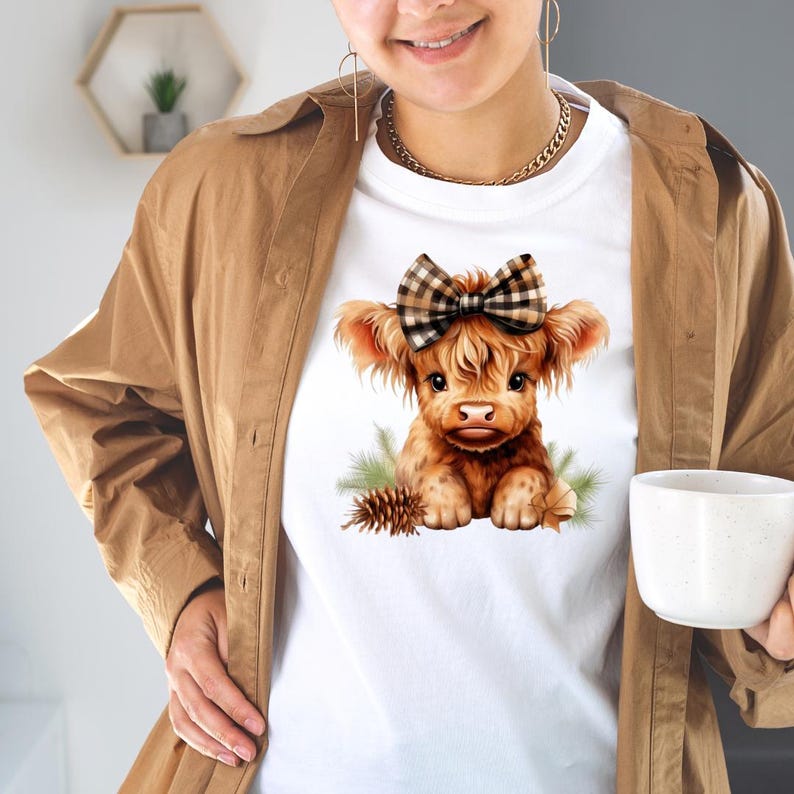 Puede incluir: Camiseta blanca con una ilustraci&oacute;n de una vaca de las Tierras Altas marr&oacute;n, con un lazo a cuadros. La vaca est&aacute; rodeada de agujas de pino y una pi&ntilde;a. La camiseta la lleva una persona con una chaqueta marr&oacute;n.