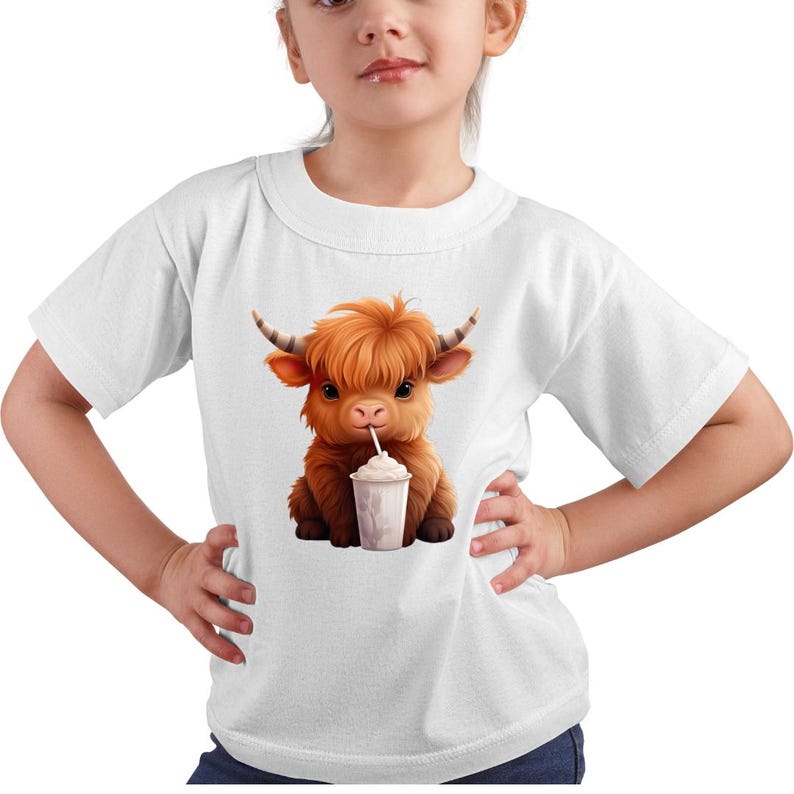 Puede incluir: Camiseta blanca con una ilustraci&oacute;n de dibujos animados de una vaca de las Tierras Altas bebiendo un batido. La vaca tiene pelaje marr&oacute;n y cuernos. El batido est&aacute; en un vaso blanco con una pajita. La camiseta tiene mangas cortas y cuello redondo.