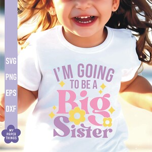 以下が含まれることがあります： ピンク、パープル、イエローの文字「I'm going to be a Big Sister」が書かれた白いTシャツ。レトロなフォントスタイルで、花と星が描かれています。