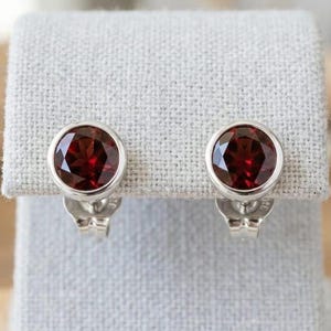 Peut inclure: Une paire de boucles d'oreilles à clous avec des pierres précieuses rondes, facettées, rouge foncé, serties dans un sertissage en argent. Les boucles d'oreilles sont présentées sur un tissu gris clair. Les pierres précieuses ont une qualité brillante et réfléchissante.
