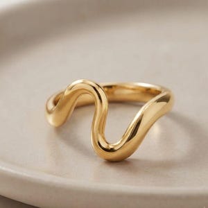 May include: A polished gold ring featuring a continuous, fluid, wavy design that curves upward and downward. The band has a smooth, reflective metallic surface with a rounded profile, creating a singular, undulating shape that encircles the finger.