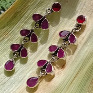 Könnte beinhalten: Silberne Hängeohrringe mit floralem Design. Jeder Ohrring hat einen kleinen, runden, roten Edelstein oben, verbunden mit drei blütenblattförmigen, tiefrosa Edelsteinen. Am unteren Ende hängt ein tropfenförmiger rosa Edelstein.