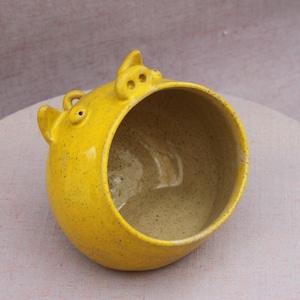 May include: A bright yellow ceramic planter shaped like a pig head with a wide, open circular top. The exterior features a speckled glaze finish, while the interior is a smooth, light beige colour. Two small ears and a snout are visible at the top.