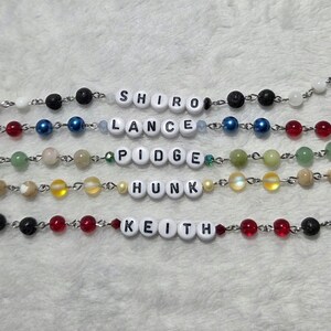 May include: Close-up of several beaded bracelets with names spelled out in white letter beads. The bracelets feature various colored beads, including black, red, blue, green, and yellow. The names include "SHIRO", "LANCE", "PIDGE", "HUNK", and "KEITH".