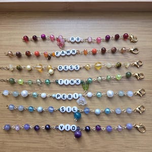 May include: A collection of beaded bracelets with gold-colored clasps. Each bracelet features a different color scheme of beads and letter beads spelling out names: RO, FAWN, DESS, TINK, PERI, SIL, and VIO. The bracelets are displayed on a wooden surface.