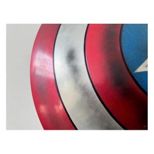 Pode incluir: Um close-up de um escudo do Capit&atilde;o Am&eacute;rica. O escudo apresenta um esquema de cores vermelho, branco e azul com um emblema de estrela. O escudo tem um acabamento met&aacute;lico e uma forma circular.