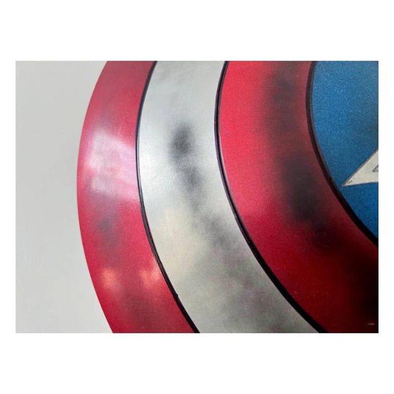 Pode incluir: Um close-up de um escudo do Capit&atilde;o Am&eacute;rica. O escudo apresenta um esquema de cores vermelho, branco e azul com um emblema de estrela. O escudo tem um acabamento met&aacute;lico e uma forma circular.