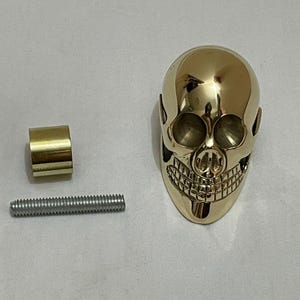 Może przedstawiać: Złoty obiekt w kształcie czaszki ze szczegółowymi rysami twarzy, cylindrycznym złotym elementem i srebrnym gwintowanym prętem. Czaszka ma wypolerowaną, odblaskową powierzchnię. Elementy wydają się być oddzielnymi częściami większego przedmiotu.