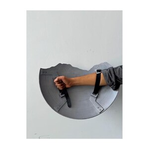 Pode incluir: Um escudo cinzento, desgastado pela batalha, com uma pega e correias pretas. O escudo &eacute; segurado pelo bra&ccedil;o de uma pessoa, mostrando o seu design e desgaste. O escudo tem uma forma semicircular com uma borda superior irregular.