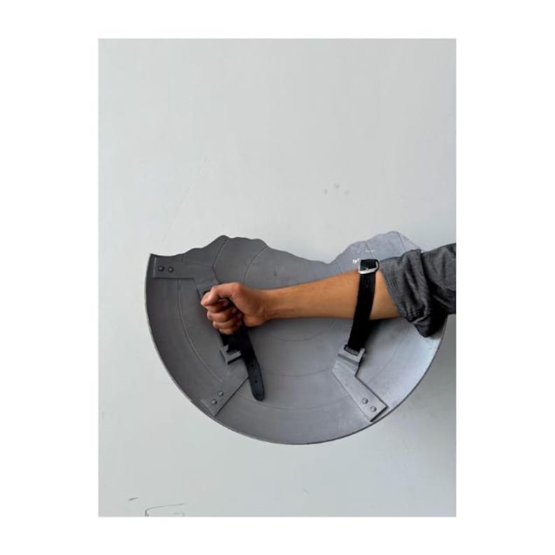 Pode incluir: Um escudo cinzento, desgastado pela batalha, com uma pega e correias pretas. O escudo &eacute; segurado pelo bra&ccedil;o de uma pessoa, mostrando o seu design e desgaste. O escudo tem uma forma semicircular com uma borda superior irregular.