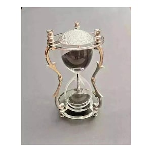 May include: An ornate hourglass with a silver-coloured metal frame and clear glass. The top and bottom are circular, and the hourglass contains dark sand. The design includes decorative flourishes and a textured top.