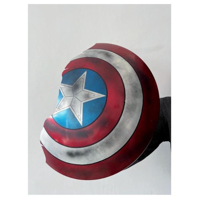 Pode incluir: Um escudo do Capit&atilde;o Am&eacute;rica com um acabamento desgastado. O escudo apresenta um centro azul com uma estrela branca, rodeado por c&iacute;rculos conc&ecirc;ntricos vermelhos e brancos. O escudo parece ser de metal e tem um aspeto desgastado e danificado.