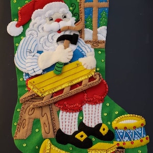 Op de afbeelding: Een groene kerstsok van vilt met een kerstmanfiguur die een hamer vasthoudt en aan een houten speelgoed werkt. De sok is versierd met pailletten en bevat een speelgoedtrein, een speelgoedtrommel en een speelgoedtrompet.