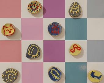 Könnte beinhalten: Eine Sammlung kleiner, handbemalter Keramikstücke mit verschiedenen Designs, darunter abstrakte Formen und Tiermotive. Die Stücke sind auf einem farbenfrohen Schachbrettmuster-Hintergrund mit rosa, lila, blauen und weißen Quadraten angeordnet. Jedes Stück ist einzigartig in Farbe und Design.