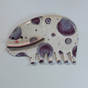 May include: A whimsical ceramic wall hanging in the shape of a creature with a white base, adorned with purple spots and speckles. It features a black and red smile, and small, stubby legs. The art piece is mounted on a white wall.