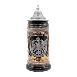 May include: A tall ceramic beer stein with a black body, featuring a metallic silver eagle emblem and the word GERMANY embossed on the front. It includes a conical pewter lid with intricate relief carvings and gold-toned decorative accents.