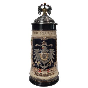 May include: A ceramic beer stein with a dark blue body, featuring a detailed emblem and text. The stein has a metal lid and a decorative eagle on top. The base and rim are black, and the overall design is ornate.