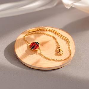 May include: A gold-coloured bracelet featuring a ladybug charm. The ladybug is red with black spots and a black head. The bracelet rests on a small, round wooden surface.