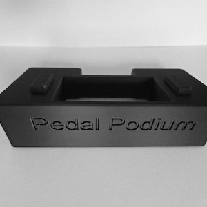 May include: Black Pedal Podium with the product name embossed on the front. The podium has a rectangular shape with a recessed centre and two raised rectangular sections on top. Designed to hold a pedal.