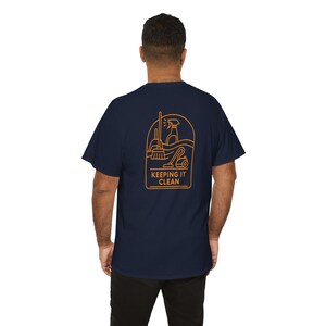 以下が含まれることがあります： オレンジ色のグラフィックが背面に施されたネイビーブルーのTシャツ。掃除道具と「KEEPING IT CLEAN」の文字が描かれています。デザインにはほうき、スプレーボトル、掃除機が含まれています。