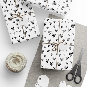 May include: White wrapping paper with a black heart pattern, tied with twine and a white heart-shaped tag. A ball of twine, scissors, and blank tags are also present, suggesting gift wrapping.